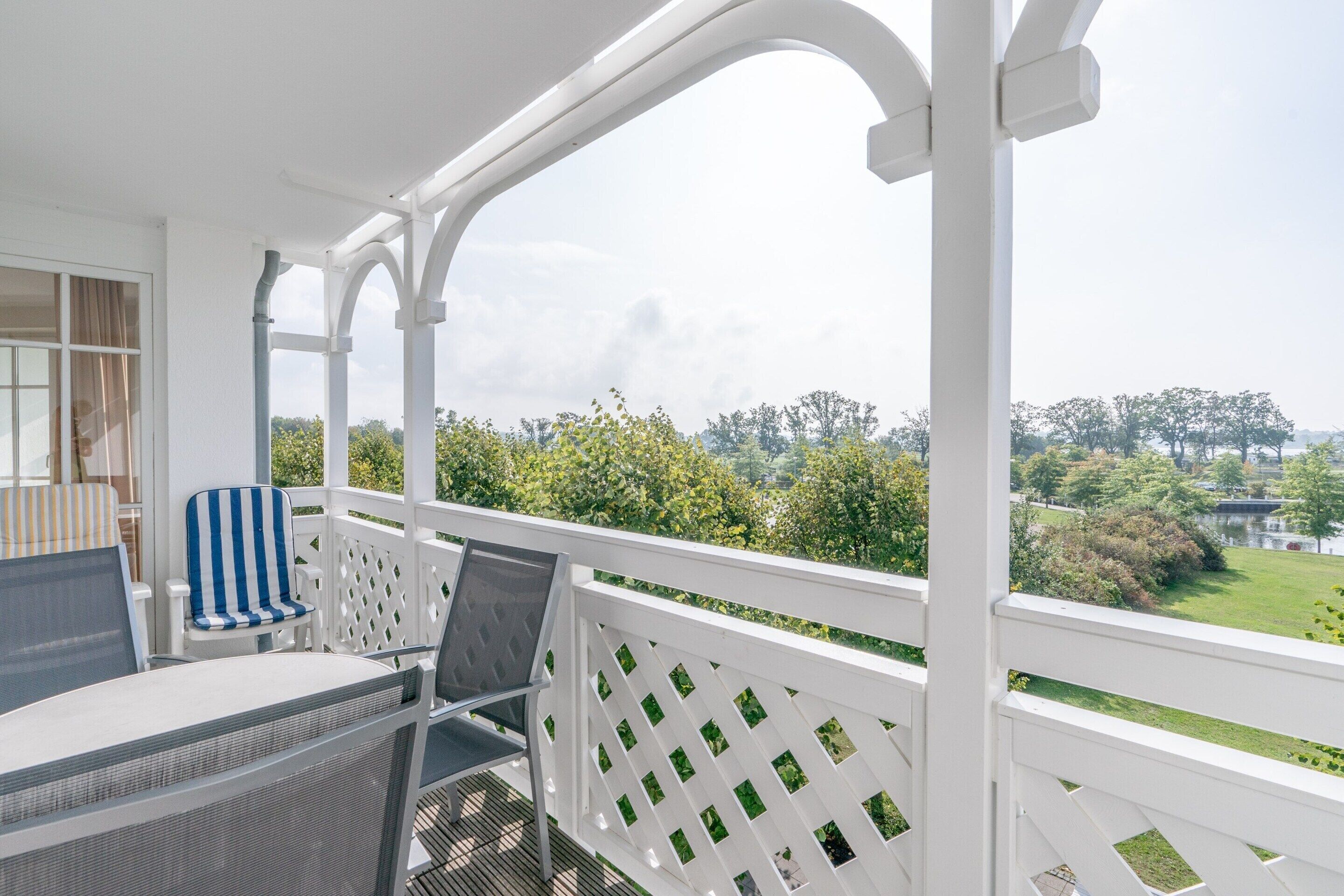 A balcony with a white lattice railing, a table with two chairs, and a view of a green landscape with trees and a body of water.