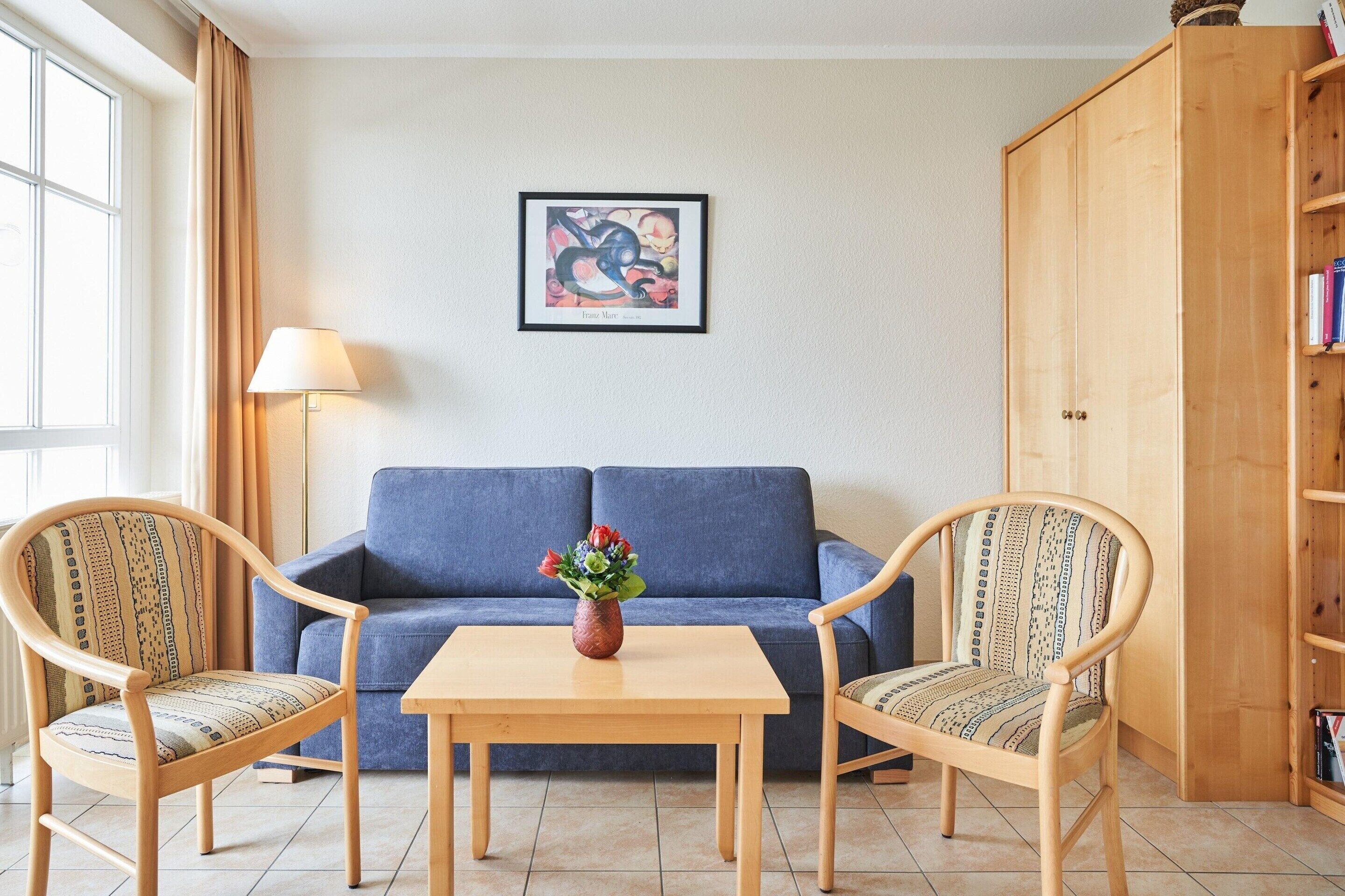 A room with a blue sofa, two patterned chairs, a wooden table with a vase, a lamp, and a wooden cabinet with books.