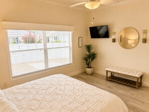 A bedroom with a large window, a bed, a potted plant, a bench, a TV, and wall sconces.