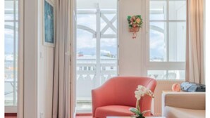 A room with a red chair, a white sofa, a vase with flowers, and a window with a view of a white fence and a ship. - Ferienwohnung im 2.og mit Balkon in Sellin (Sellin)