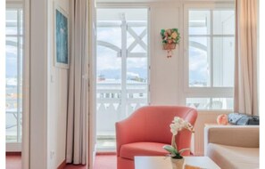 Ein Zimmer mit einem roten Sessel, einem weißen Sofa, einer Blumenvase und einem Fenster mit Blick auf einen weißen Zaun und ein Schiff.