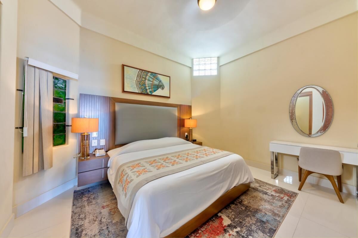 A hotel room with a bed, bedside lamps, a desk, a chair, a mirror, and a window with curtains.