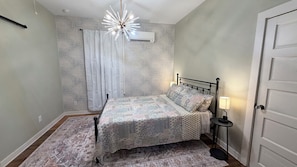 A bedroom with a bed, a nightstand, a lamp, an air conditioner, and a patterned wall. - Pleasant 2-bedroom, 2 bathroom house in charming Galveston. (Galveston)