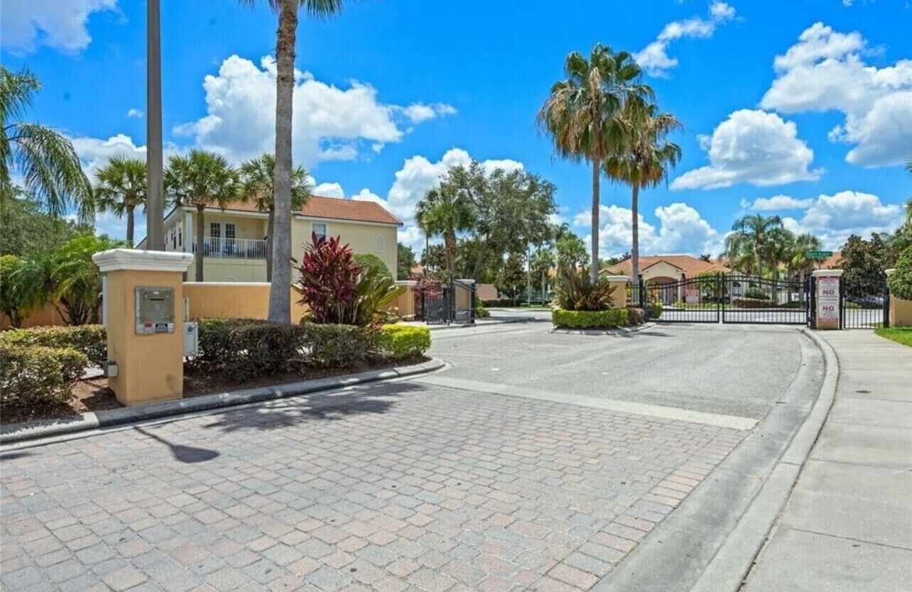 A gated residential area with a paved driveway, palm trees, and a clear blue sky.