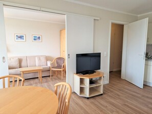 A living room with a sofa, chairs, a TV on a stand, a dining table, and a kitchen in the background. - Ferienwohnung mit Hund in Sellin (Sellin)