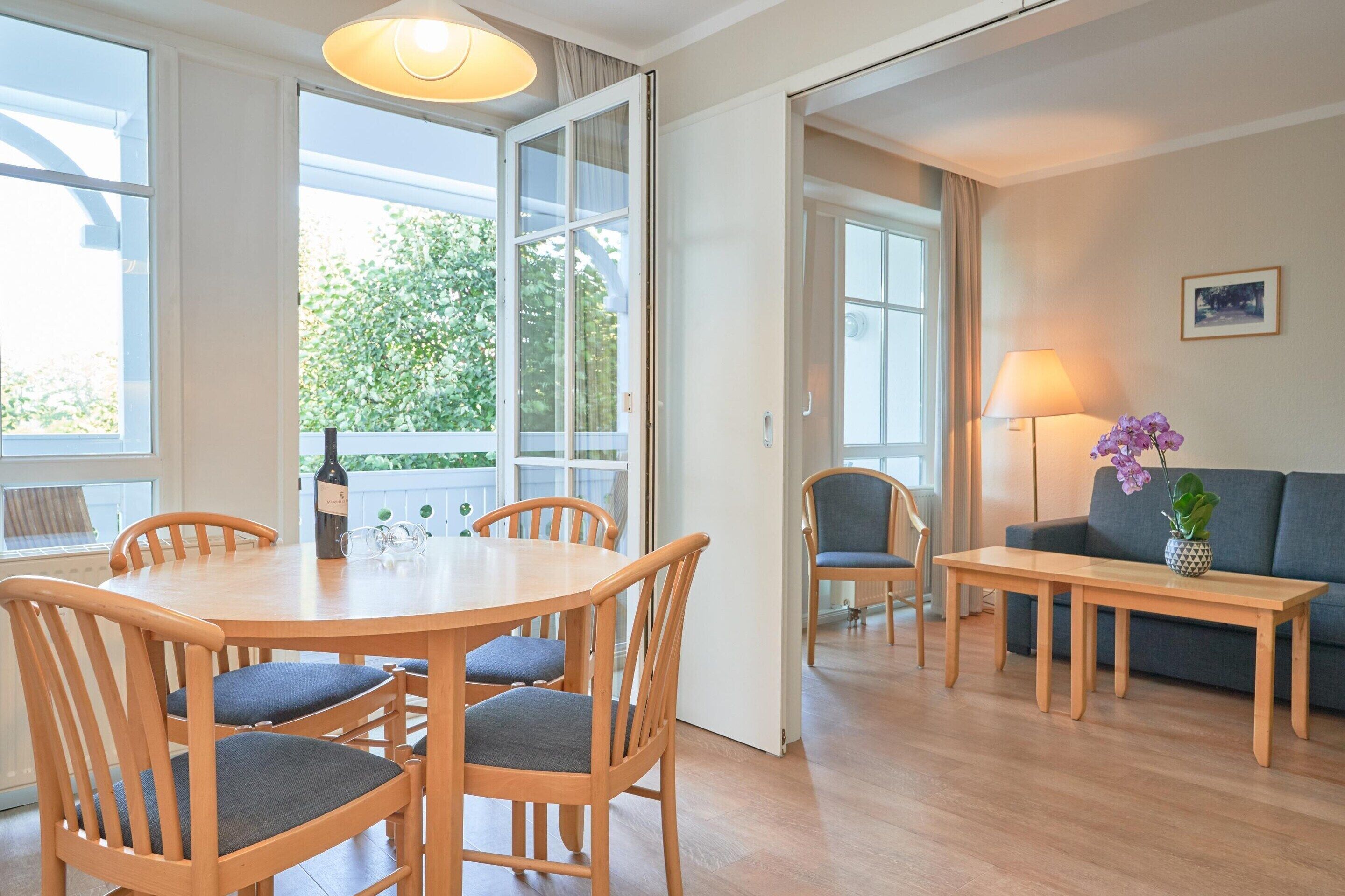 A dining area with a wooden table and chairs, a sofa, a lamp, and a view of greenery outside.