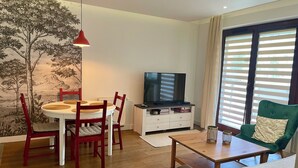 Uma sala de estar com uma mesa de jantar, cadeiras, uma TV, um sofá e uma janela com persianas.