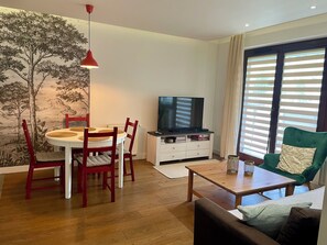 Een woonkamer met een eettafel, stoelen, een televisie, een bank en een raam met jaloezieën.