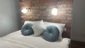 Cama com colcha branca e duas almofadas azuis, diante de uma parede de tijolos com duas luzes fixadas.