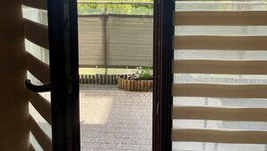 Vista pela janela com persianas horizontais, área externa pavimentada e um edifício ao fundo.