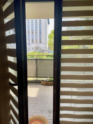 Uitzicht door een raam met horizontale jaloezieën, een geplaveid buitengebied en een gebouw op de achtergrond.
