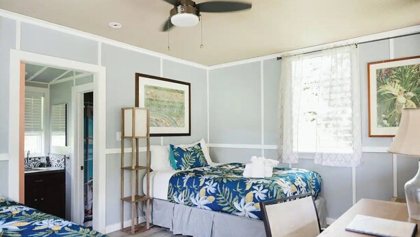 A bedroom with two beds, a ceiling fan, a window with curtains, a bedside table, and a framed picture on the wall.