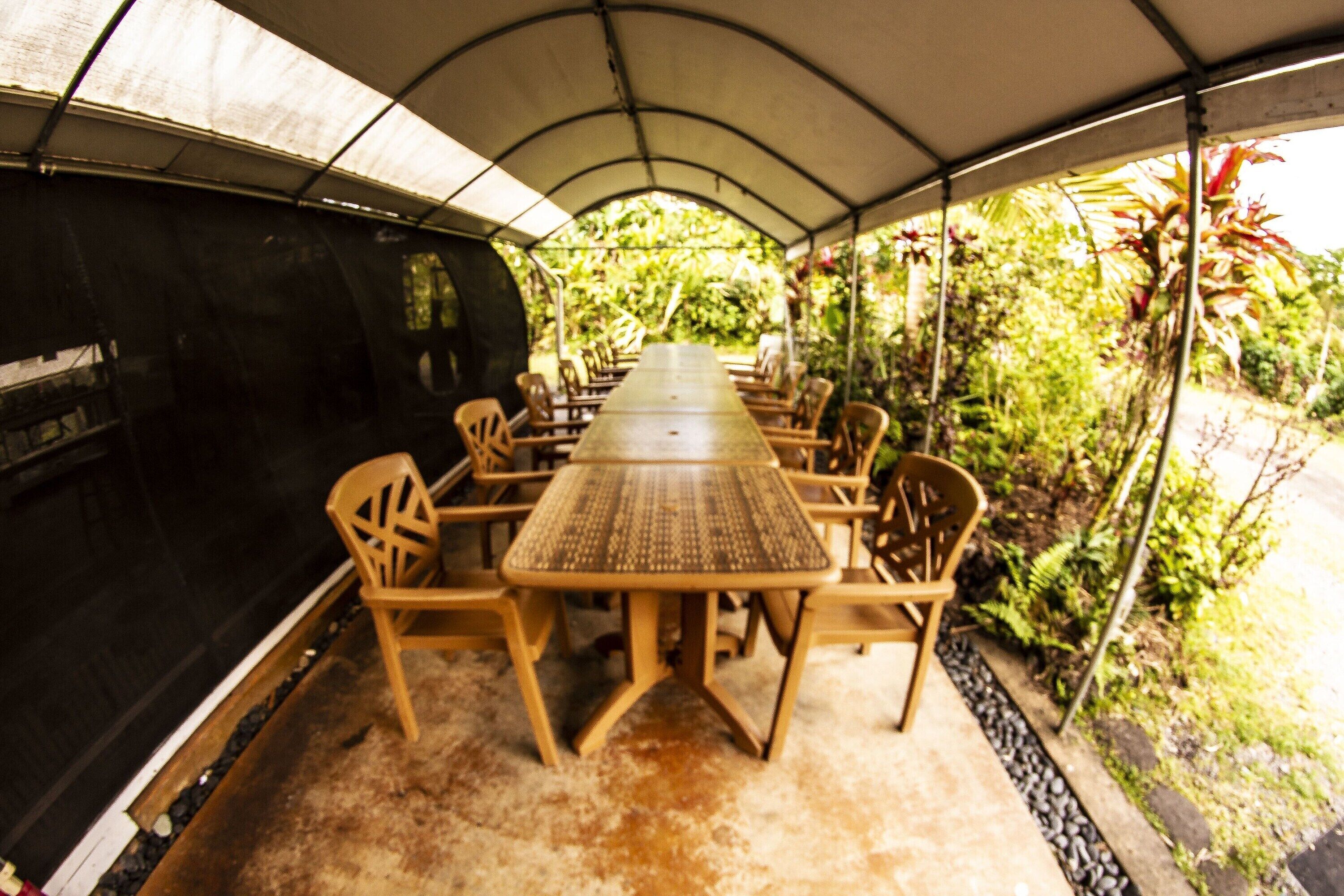 A covered outdoor dining area with a long table and chairs, surrounded by greenery.