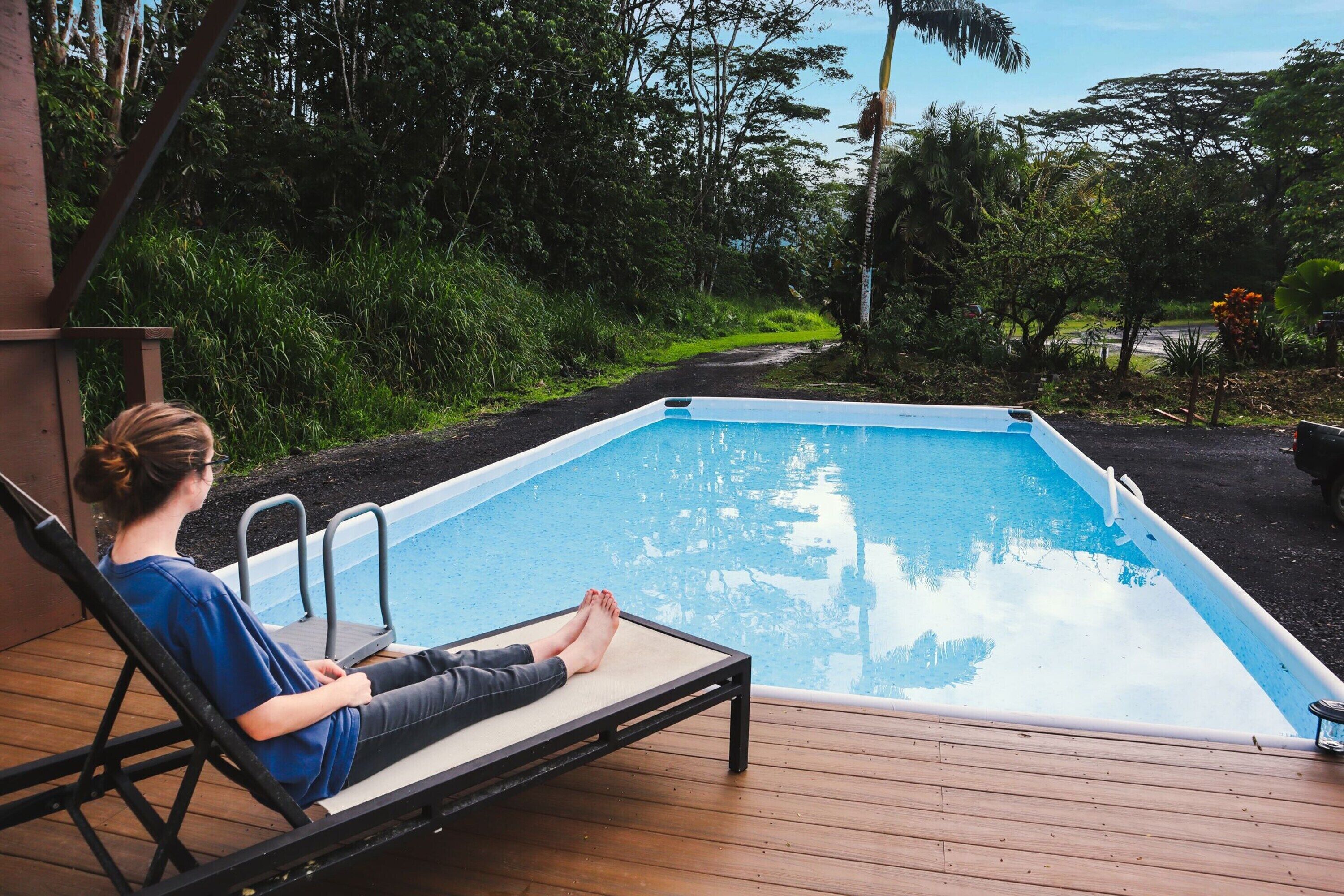 A person relaxing on a lounge chair by a swimming pool with a view of a lush garden and a road in the background.