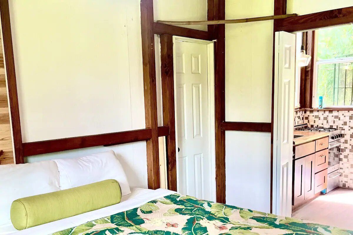 Una habitación con cama, almohada verde, techo de madera y vista a una cocina con salpicadero de azulejos.