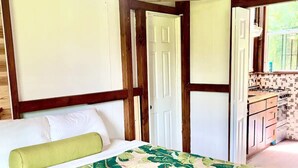 A room with a bed, a green pillow, a wooden ceiling, and a view of a kitchen with a tiled backsplash.