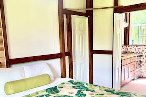 Ein Zimmer mit einem Bett, einem grünen Kopfkissen, einer Holzbalkendecke und Blick auf eine Küche mit gefliestem Spritzschutz.