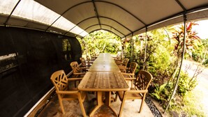 A covered outdoor dining area with a long table and chairs, surrounded by greenery.