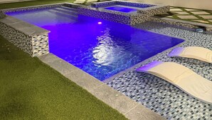 Une piscine dont le bord est recouvert de carreaux et qui est agrémentée d’un éclairage bleu, entourée de gazon synthétique et d’une clôture blanche.