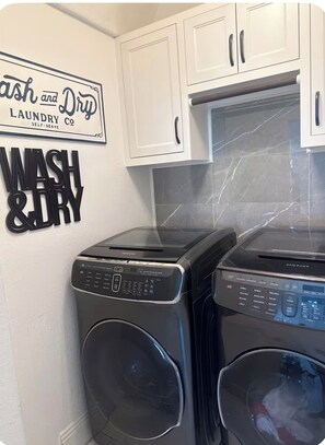 Un espace buanderie équipé de deux machines à laver à chargement frontal, d’un panneau mural indiquant « Wash and Dry Laundry Co. – laverie en libre-service », et de placards blancs au-dessus des machines.
