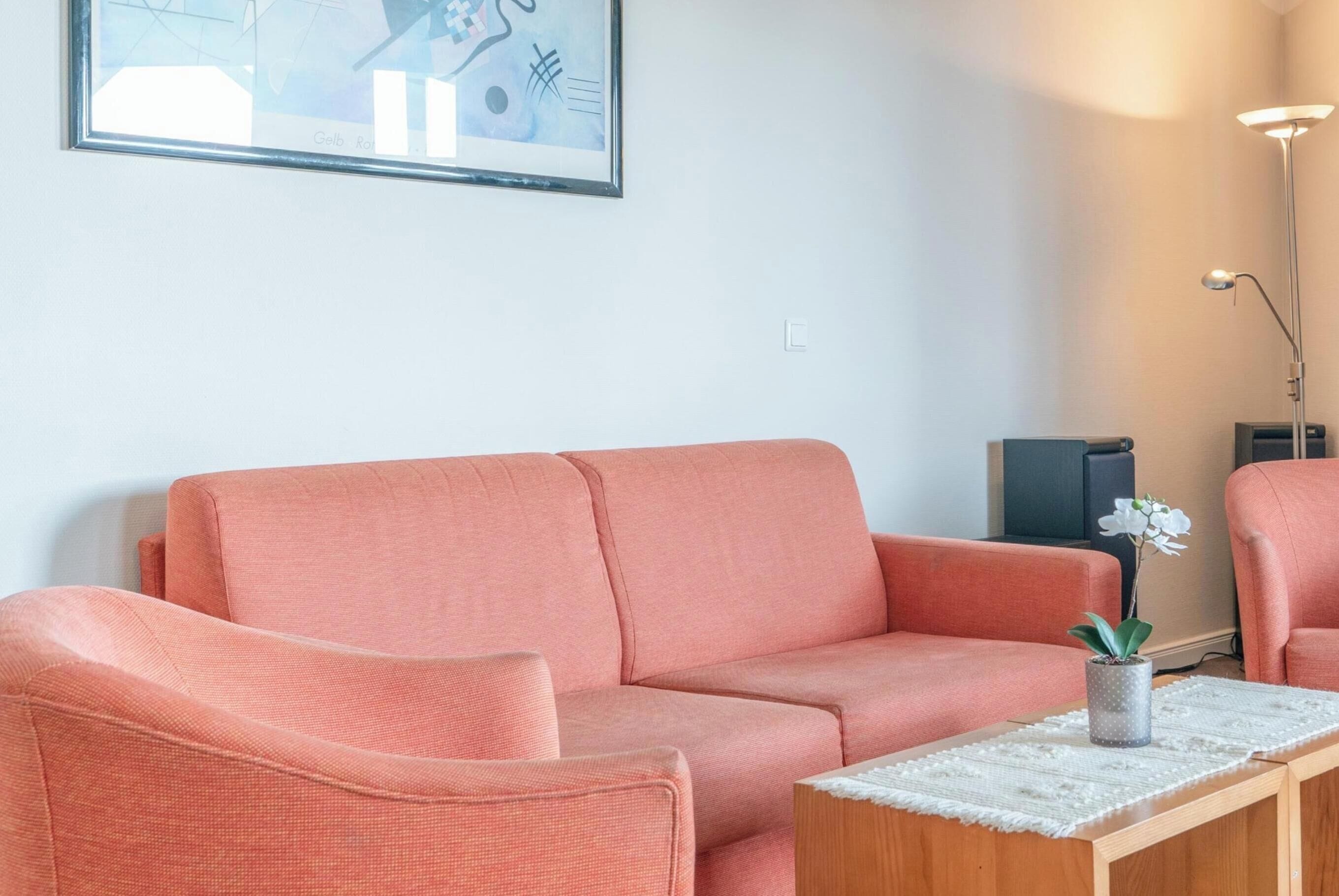 Una sala de estar con un sofá y un sillón, una mesita con un florero, y un cuadro abstracto enmarcado en la pared.