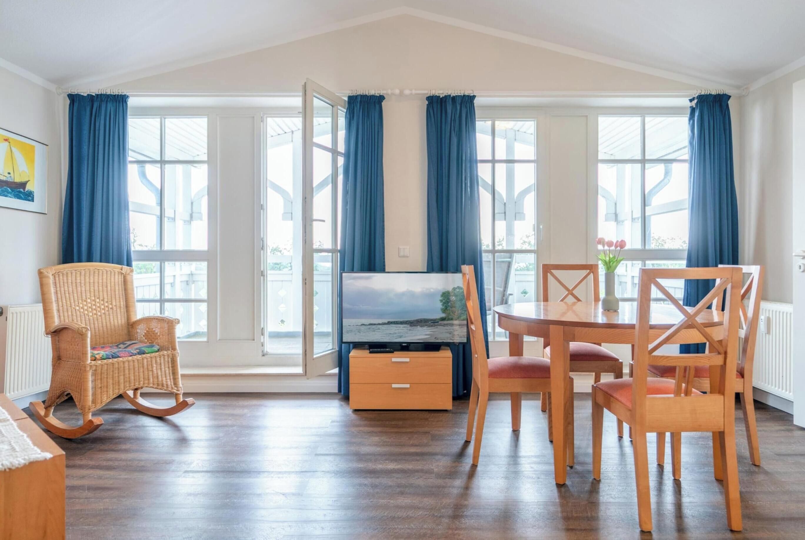 A room with a television, a dining table with chairs, a rocking chair, and large windows with curtains.