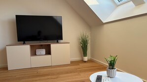 A modern living room with a flat-screen TV on a wooden stand, a white console table, a potted plant, a small white table with remote controls, and a skylight.