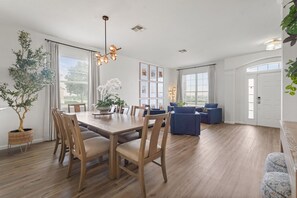 Ein geräumiger Essbereich mit einem Holztisch und Stühlen, einer Chandelieber und einem großen Fenster mit Vorhängen.
