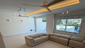 Ein modernes Wohnzimmer mit einem großen, im Winkel angeordneten Ecksofa, einem Deckenventilator, Einbaustrahlern und einem großen Fenster mit Außenblick.