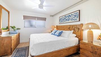 A bedroom with a wooden bed, a bedside table, a lamp, a window with blinds, a ceiling fan, and a wall-mounted artwork.