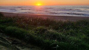 Escena de playa con el sol poniéndose sobre el océano, dunas cubiertas de hierba en primer plano y un cielo despejado.