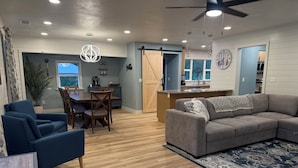 Un espacio habitable amplio con un sofá gris, sillones azules, una mesa de comedor y una cocina al fondo.