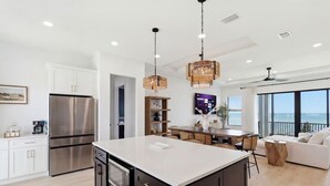 A modern kitchen with a large island, stainless steel appliances, and a dining area with a view. - Sunset Oasis: Beachfront Bliss with 2 Pools, Fire Pits & Room for 16 (Melbourne Beach)