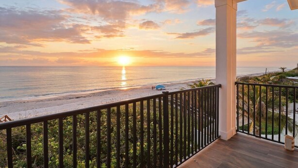 A balcony with a view of the ocean at sunset, a railing, and a wooden floor. - Sunset Oasis: Beachfront Bliss with 2 Pools, Fire Pits & Room for 16 (Melbourne Beach)