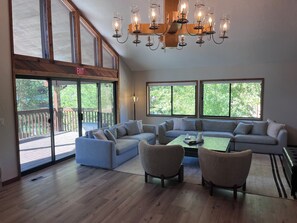 Amplia sala de estar con un sofá modular grande, dos sillones, una mesa de centro y un candelabro. Amplias ventanas ofrecen luz natural y vistas al exterior.