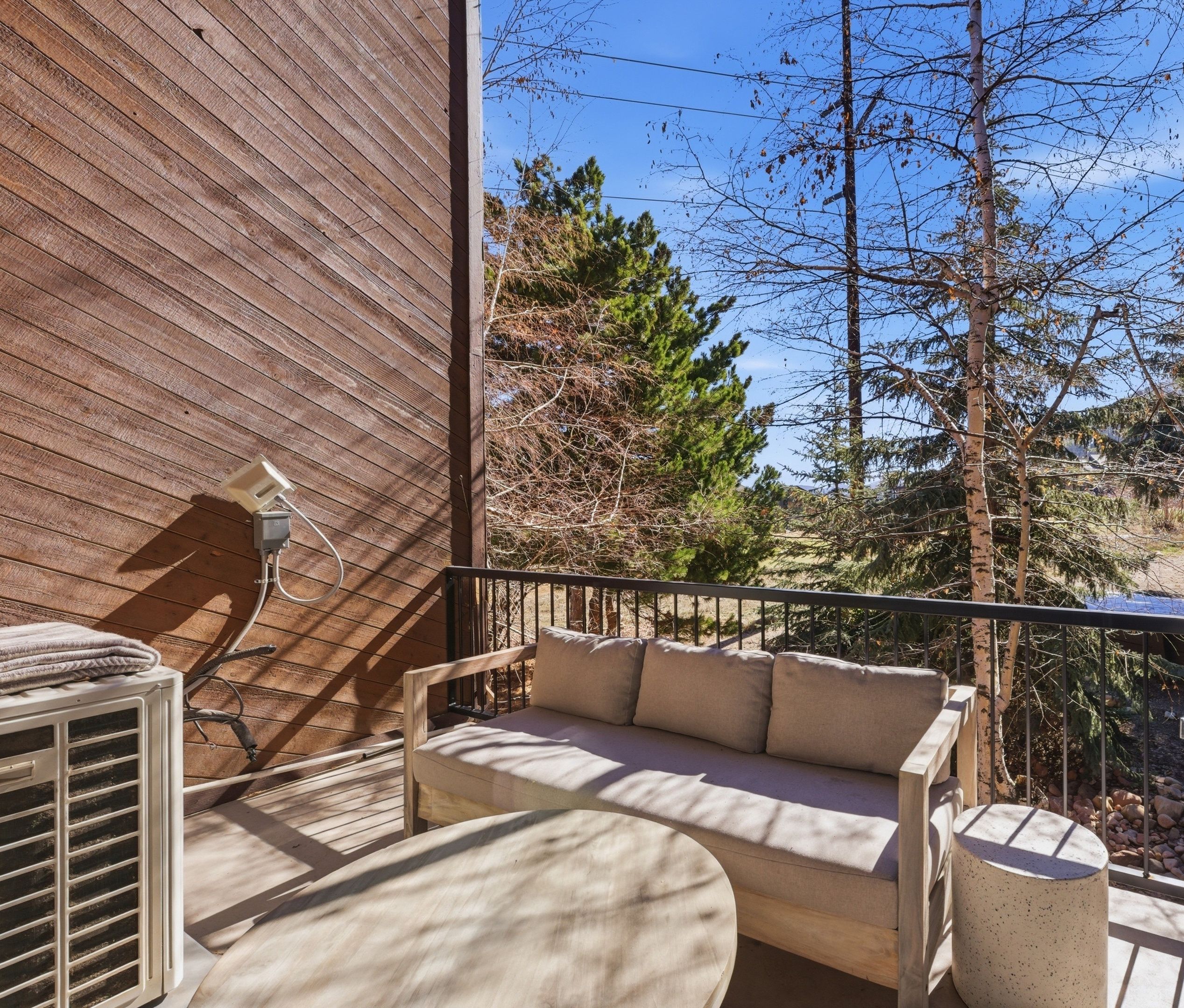 A wooden deck with a sofa and a small table, a power outlet, and a view of trees and a clear sky.