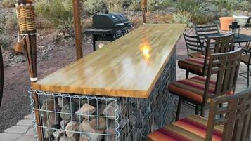 An outdoor bar area with a wooden counter, metal grill, and chairs.