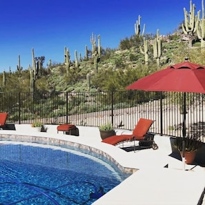 A pool area with lounge chairs and a red umbrella, surrounded by a fence and cacti.