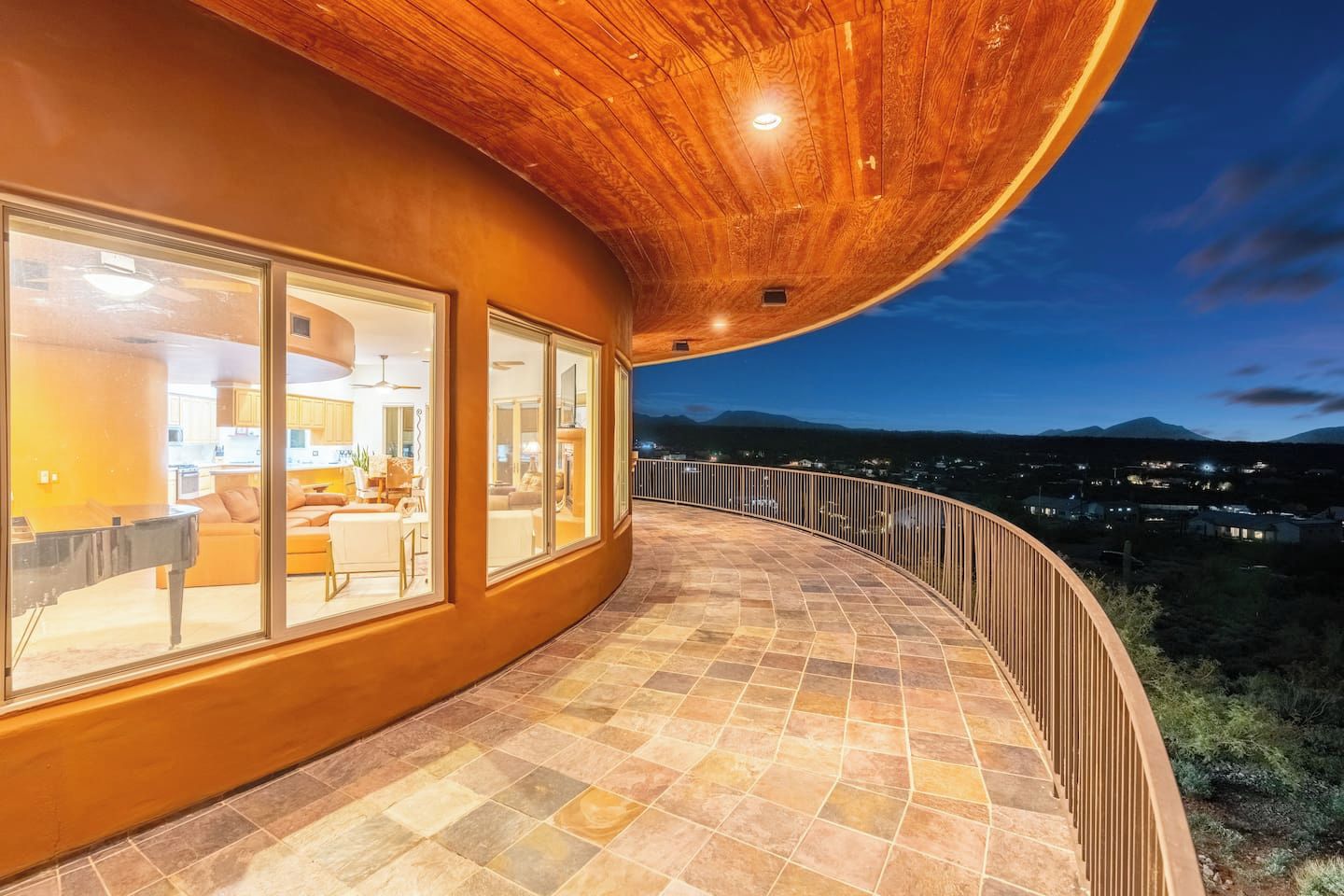 A curved balcony with a stone floor, large windows, and a view of a town at dusk.