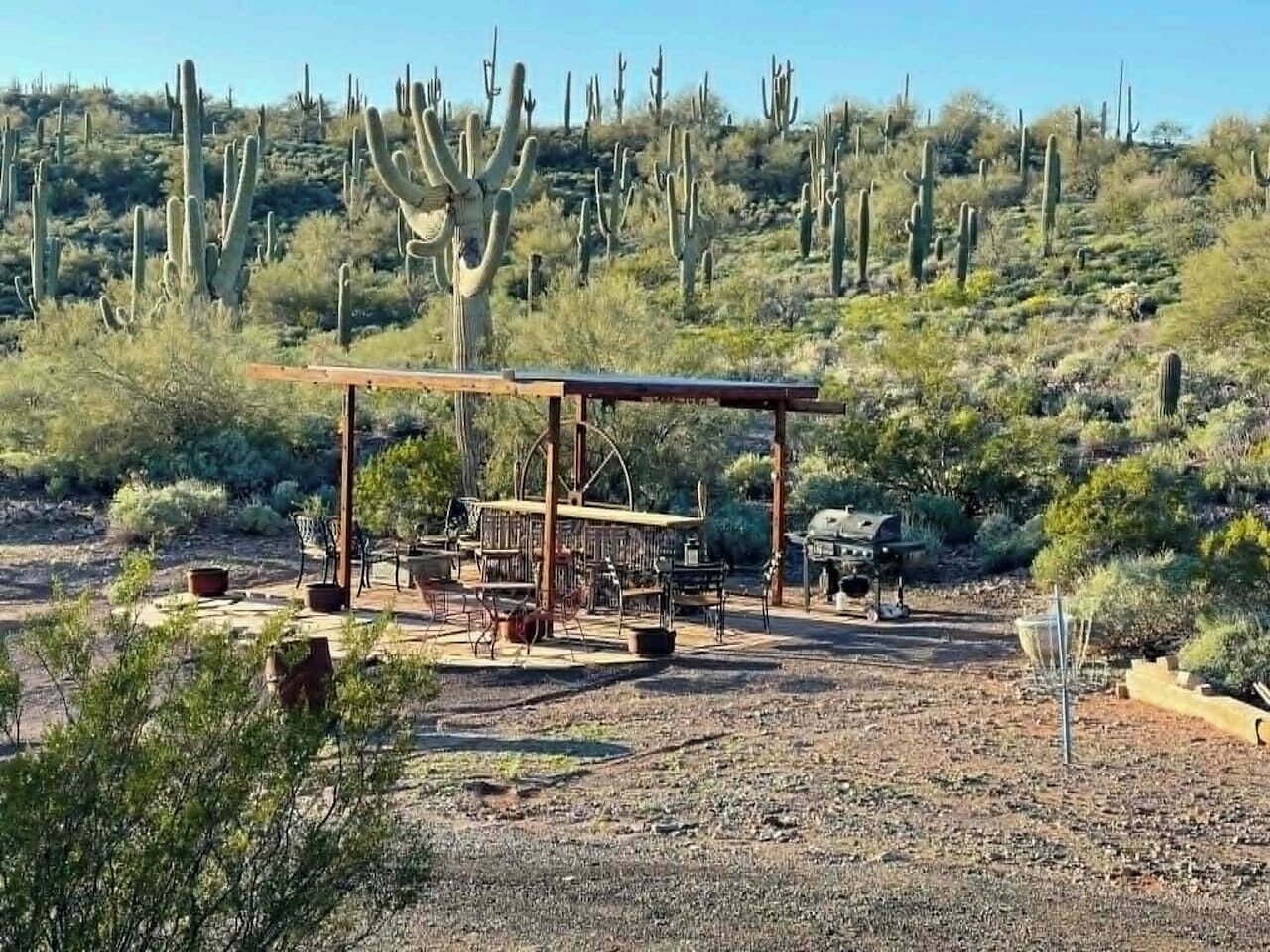 A desert landscape with cacti, a wooden structure with tables and chairs, and a grill.