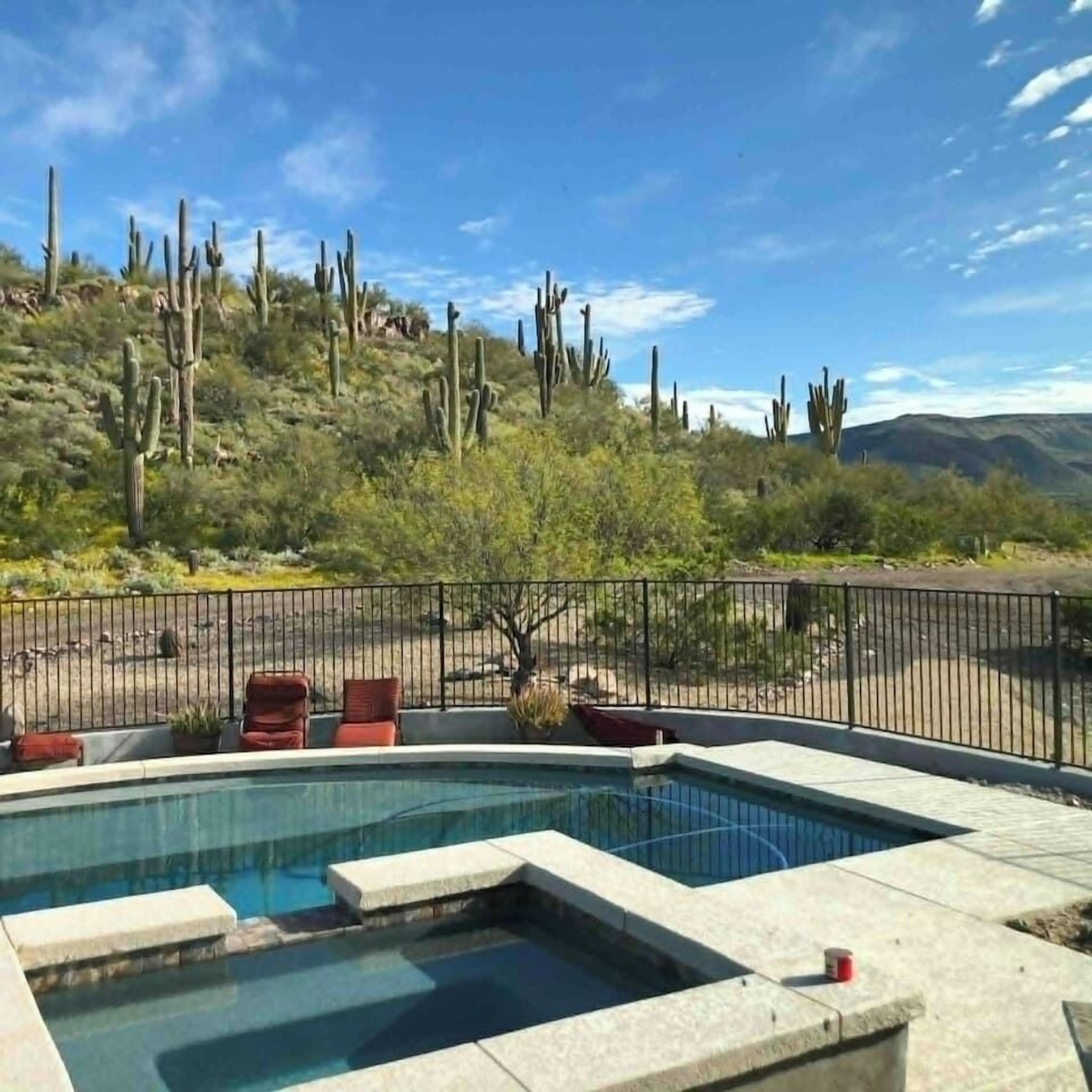 A pool with a surrounding fence, red chairs, and a desert landscape with cacti in the background.