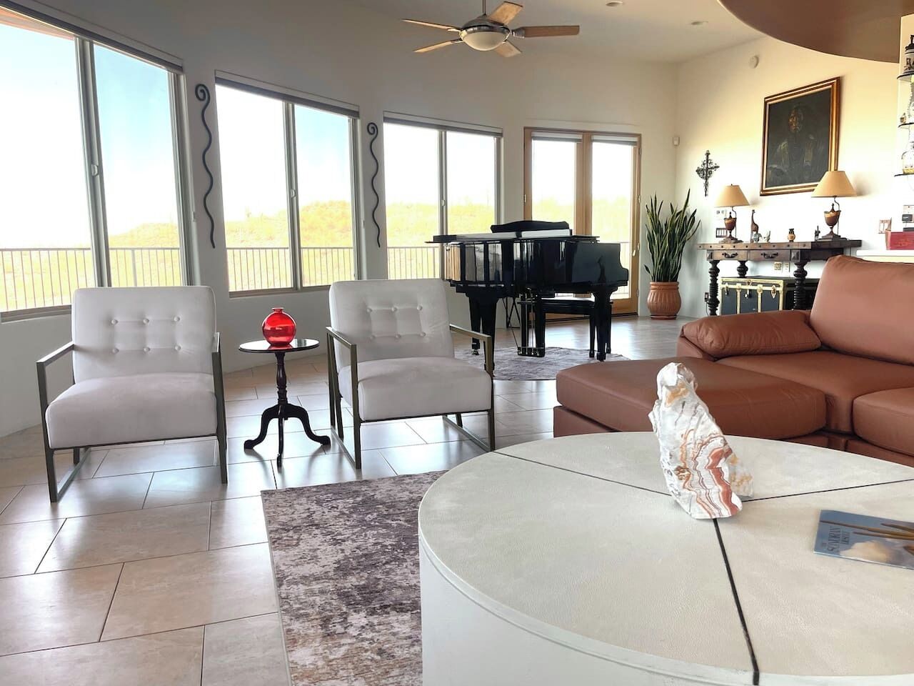A spacious living room with a piano, two armchairs, a coffee table, and a view through large windows.
