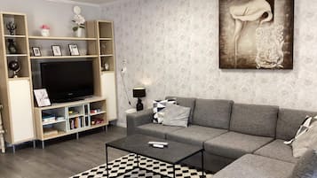 Uma sala de estar com sofá cinza, mesa de centro preta, tapete xadrez, televisão sobre um suporte de madeira e uma parede com padrão floral.
