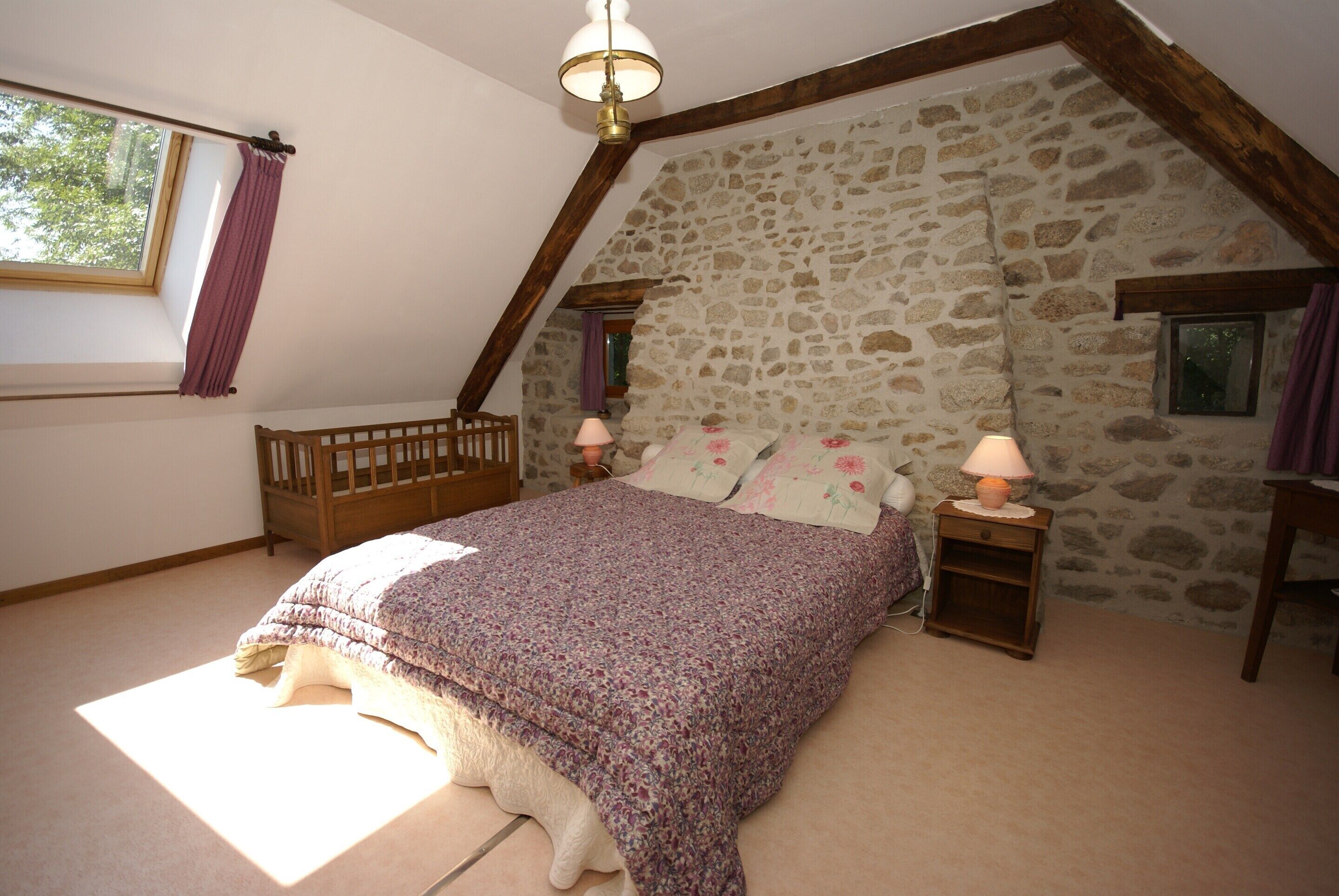 A bedroom with a stone wall, a wooden crib, a bed with a floral bedspread, two bedside tables with lamps, and a window with curtains.