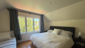 Um quarto com uma cama grande, uma cama menor, uma janela com cortinas e uma mesa de cabeceira com abajur.