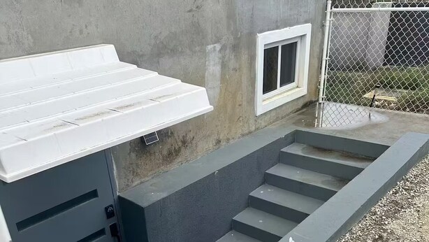 Een betonnen constructie met trappen die leiden naar een witte deur, een metalen hek en een grindpad.