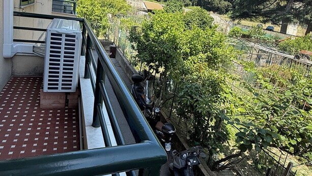 Een balkon met een betegelde vloer, een geparkeerde scooter en een rij bomen op de achtergrond.