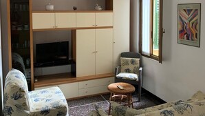 Een woonkamer met een televisie, bank, stoel en een kast met plank.