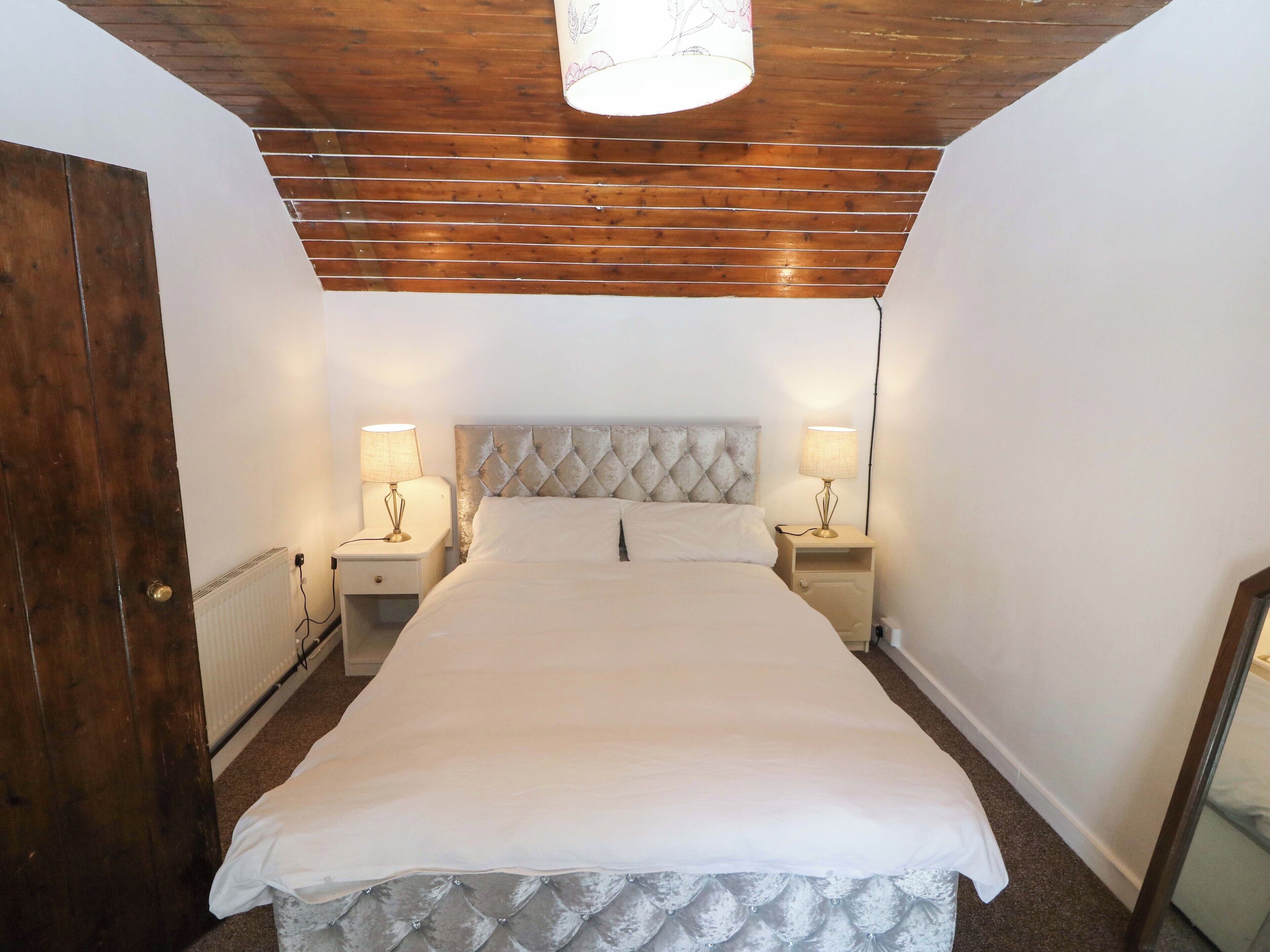 Un dormitorio con techo de madera, una cama con cabecera capitonada, dos mesitas de noche con lámparas y una puerta.