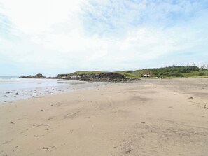 Ein Strand mit sandigem Ufer, ein Hügel mit Grasbewuchs und ein Gebäude sowie eine felsige Landzunge in der Ferne.